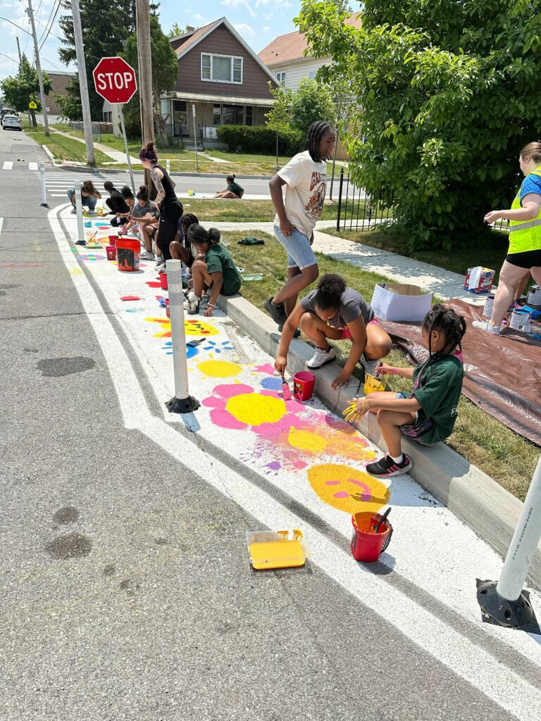 Approximately one dozen children paint brightly colored artwork, like flowers and smiley faces, within the white boundary of a protected curb extension.
