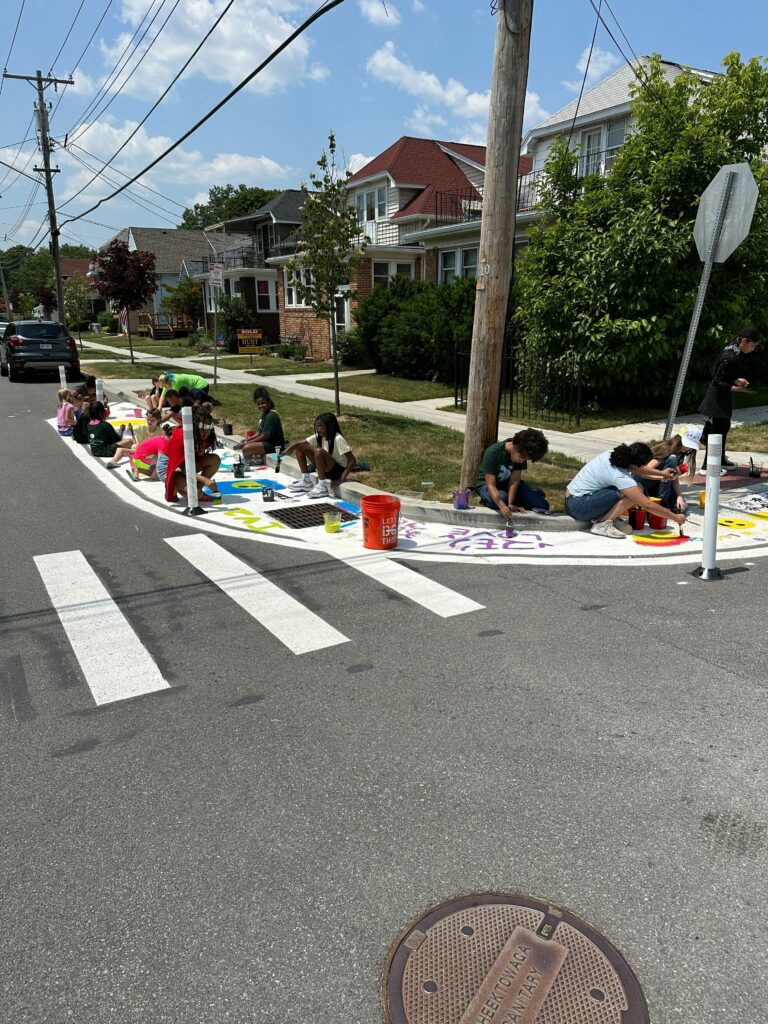 Approximately one dozen children painting artwork within the boundaries of a white curb extension, adjacent to a crosswalk.